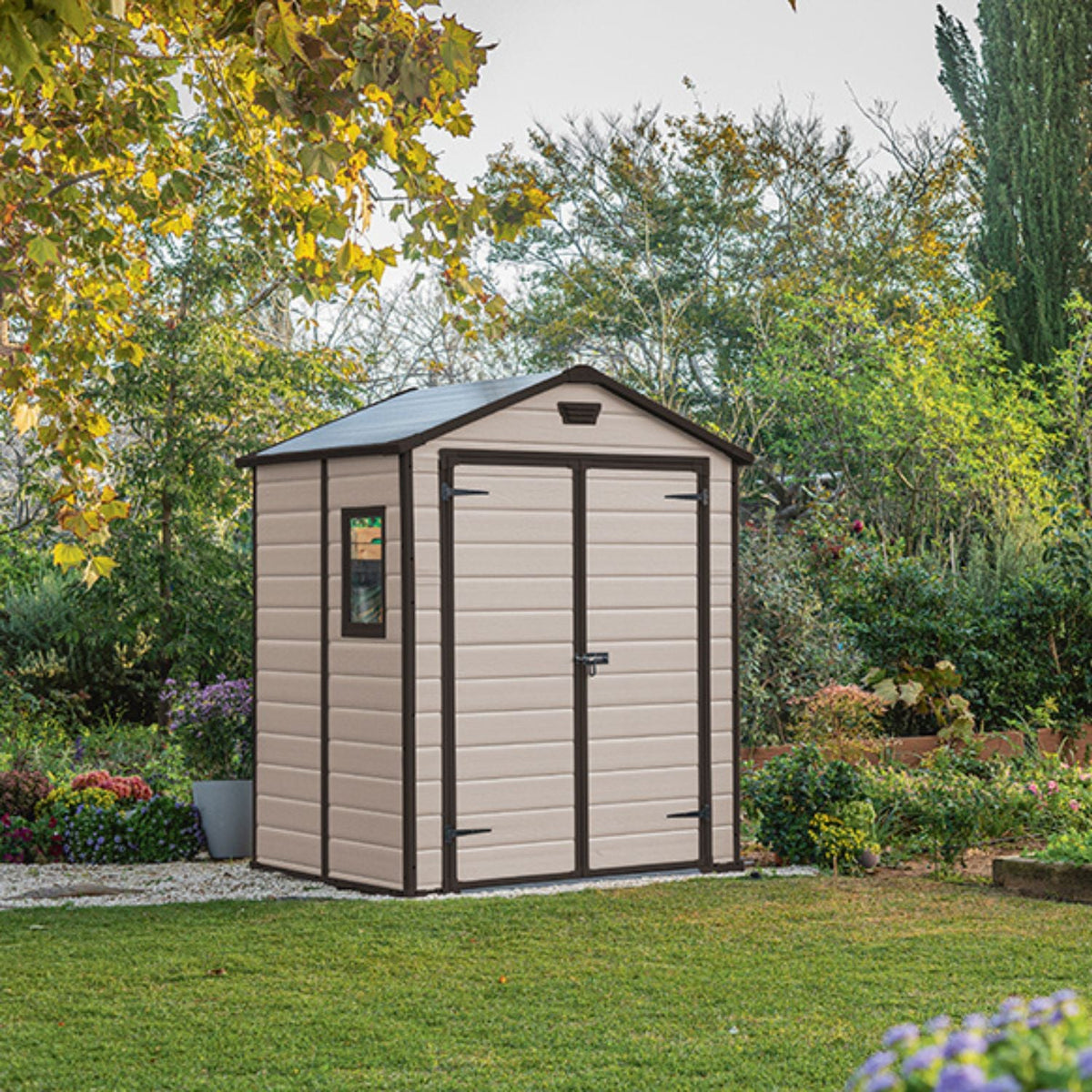 Abri de jardin MANOR beige fermé, installé sur une pelouse dans un jardin arboré