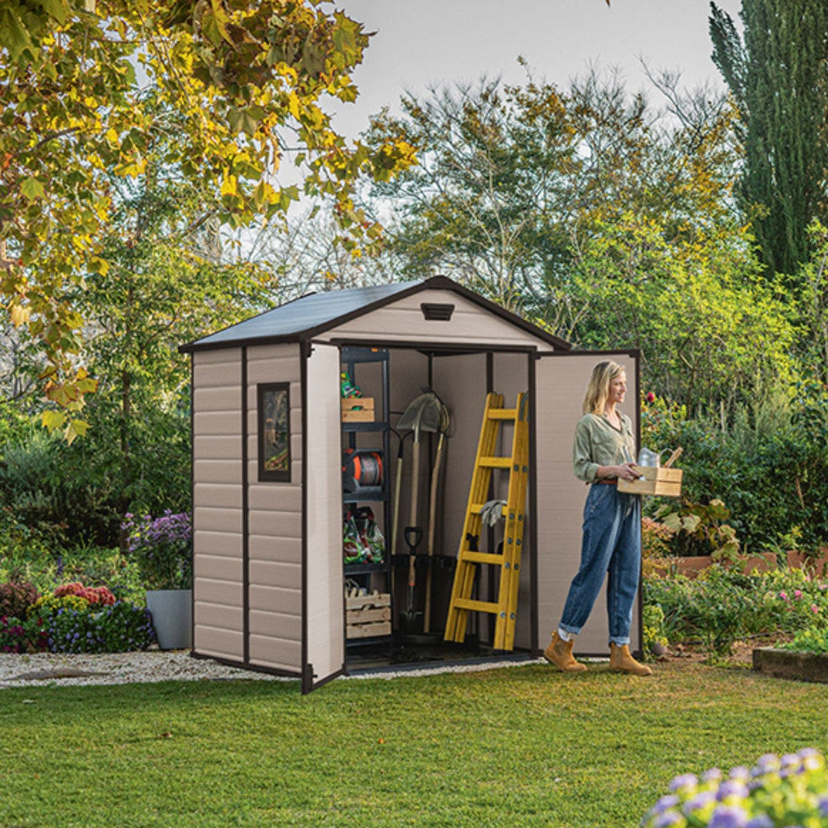 Abri de jardin MANOR beige ouvert avec outils rangés et femme portant une caisse