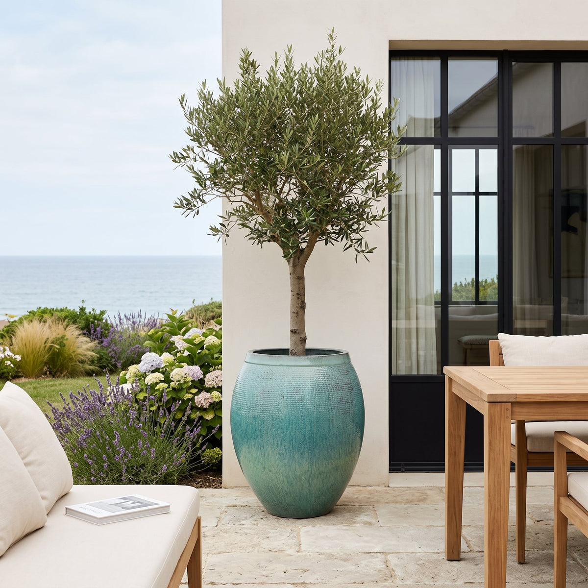 Pot Cocoon 66x76 cm bleu océan en terre cuite émaillée, olivier près d'une table en terrasse