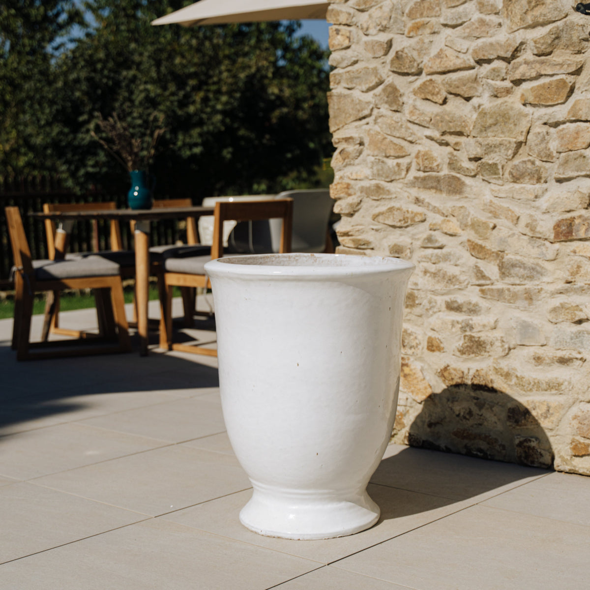Macetero Arthur con pie 42x52 cm blanco de cerámica esmaltada, en una terraza junto a un muro de piedra