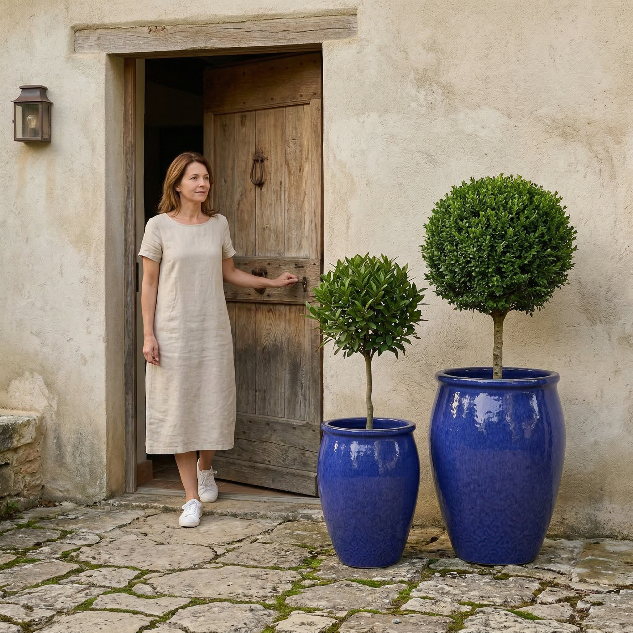 Keramický květináč BACCHUS - Majorellová modř s kulatým buxusem před starými dřevěnými dveřmi. Žena v letních bílých šatech pečuje o rostlinu.